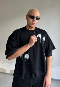 Μαύρο oversized T-shirt με μοναδικό σχέδιο λευκής πιτσιλιάς από μπογιά, κοντά μανίκια και δομικές λεπτομέρειες, επενδυμένο με διάφανη αμάνικη βάση.