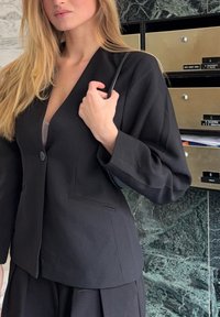 Blazer noir cintré avec un col en V profond, une fermeture à un seul bouton et deux poches avant. Le tissu semble texturé, associé à un haut clair en dessous.