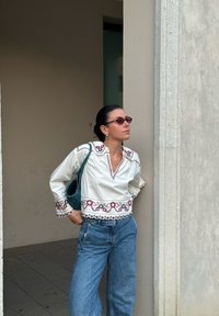 Bijela bluzica s vezom i šarenim uzorcima, upairana s svijetloplavim, širokim trapericama. Dodaci uključuju zeleni, pleteni ruksak i naočale s nijansama.