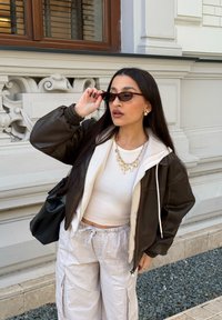 Žena upravující malé tmavé sluneční brýle, oblečená v bílé vrstvené halence, hnědé kožené bundě, širokých světle barevných kalhotách, se zlatými řetízkovými náhrdelníky, stojící venku.