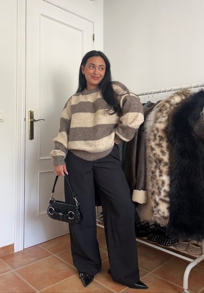 Suéter de punto de grandes dimensiones en tonos beiges y marrones con rayas, combinado con pantalones de pierna ancha negros y un pequeño bolso negro. El fondo presenta abrigos en un perchero.