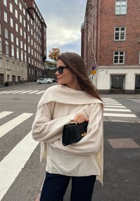 Sweater oversized de cor creme com mangas largas, combinado com calças de cor azul-marinho. Bolsa de mão preta com fecho metálico. Fundo urbano.