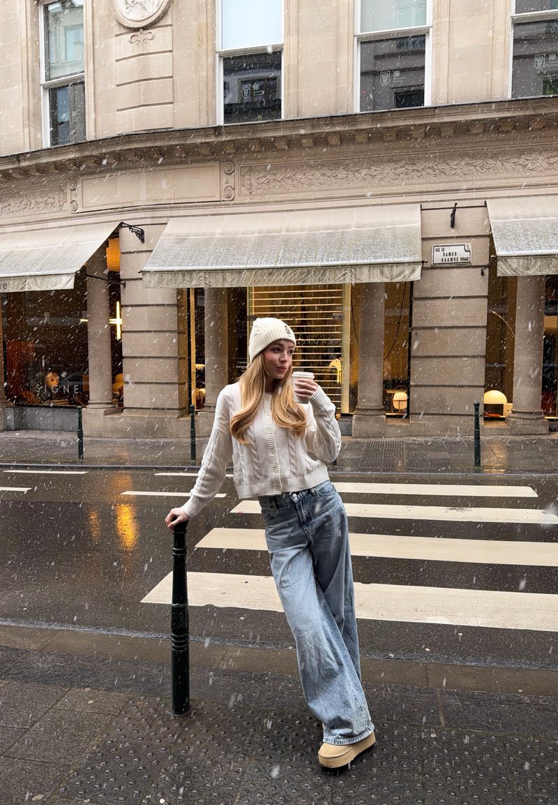 Ung kvinna i vit hatt och tröja, vida jeans, dricker kaffe medan hon lutar sig mot en gatstolpe nära ett övergångsställe i lätt snöfall.