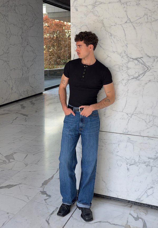 Joven con cabello rizado que lleva una camiseta negra ajustada y vaqueros azules, de pie junto a una pared de mármol blanco en el interior.