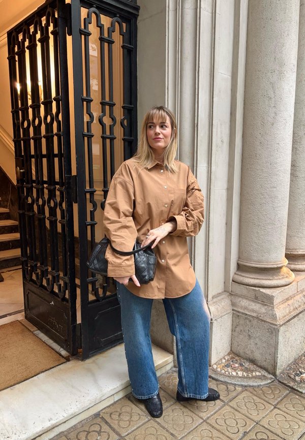 Femme debout devant un bâtiment avec de grandes colonnes en pierre, portant une chemise marron oversize, un jean bleu, et tenant un sac à main noir matelassé.