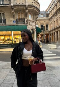 Svart zip-up jacka, vit crop-top med logotyp, svarta byxor, håller en genomskinlig kopp med dryck, och en liten röd handväska. Utomhusscen på en gata.