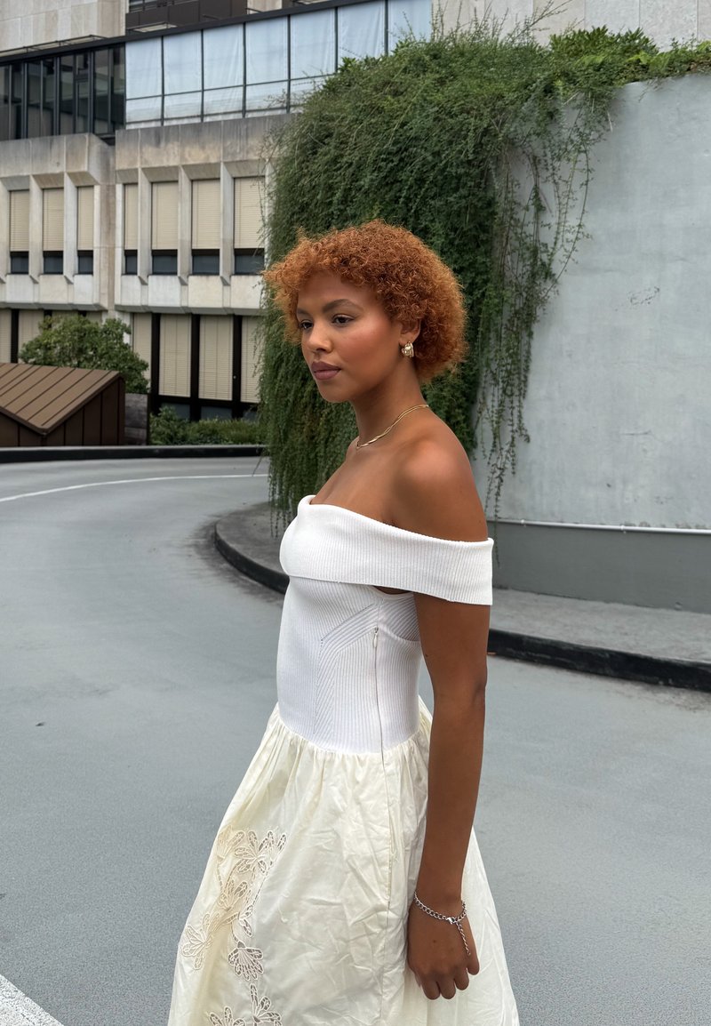 Eine Frau mit lockigem roten Haar, die ein schulterfreies weißes Kleid trägt, steht auf einer gebogenen städtischen Straße mit grüner Vegetation und Gebäuden im Hintergrund.
