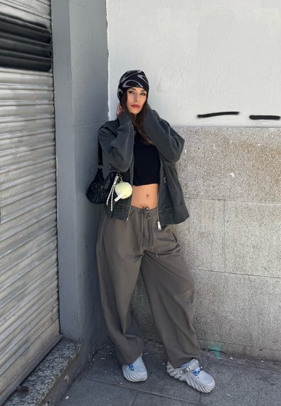 Joven mujer con pantalones grises holgados, top negro corto, chaqueta oversize, gorro con estampado y zapatos blancos gruesos está apoyada contra una pared urbana.