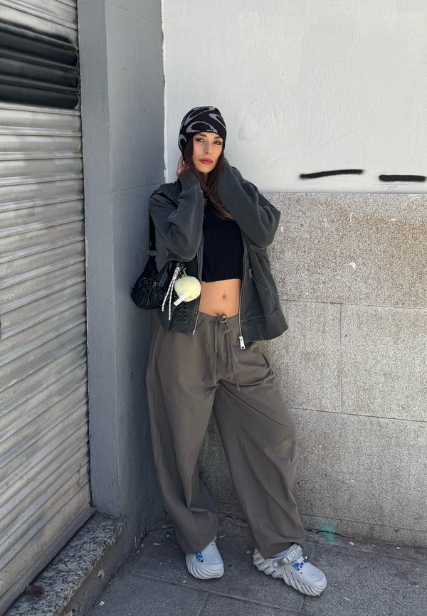 Junge Frau in locker sitzender grauer Hose, schwarzem cropped Top, übergroßer Jacke, gemusterter Mütze und weißen klobigen Schuhen steht an einer urbanen Wand.