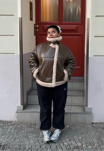 Chaqueta de cuero marrón con ribete de borreguito beige, cierre de cremallera y bolsillos laterales, combinada con pantalones deportivos negros y zapatillas plateadas.