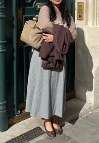 Grau fließender Rock; beige Pullover mit strukturiertem Muster; braune Jacke über dem Arm; beiger Handtasche; braune Schuhe mit Akzenten; stehend neben einer Tür.