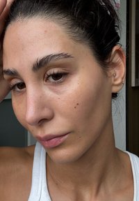 Nærbilde av et ansikt med lys hud, naturlig sminke og brune øyne. Håret er mørkt, lett bølgete; iført små gulløreringer. Bomullstank topp synlig.