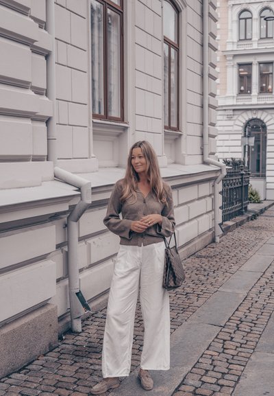 Cárdigan marrón de botones, pantalones de pierna ancha blancos, zapatos beige, bolso estampado. Pavimento de piedra y edificio blanco de fondo.