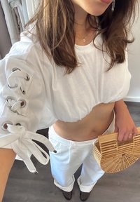 Λευκή κοντή μπλούζα με κορδόνια στα μανίκια, που συνδυάζεται με λευκά παντελόνια με φαρδιά γραμμή. Το αξεσουάρ είναι μια τσάντα από μπαμπού, που προσθέτει υφή στη σύνθεση.