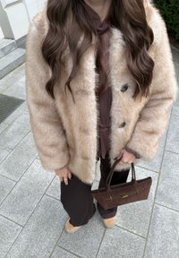 Femme aux longs cheveux bruns ondulés portant un manteau en fausse fourrure beige, un chemisier marron, un pantalon noir, des chaussures camel et tenant un sac à main marron foncé.