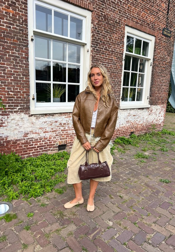 Rudas odinė trumpa striukė, balta viršutinė dalis, beige sijonas ir šviesūs butai. Rudas rankinės su metaliniais akcentais. Raudonų plytų siena ir žaliuojančių augalų fono.
