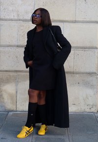 Schwarzer Mantel über einem schwarzen Kleid, mit schwarzen kniehohen Socken und gelben Sportschuhen mit schwarzen Akzenten. Steht vor einer Steinmauer.