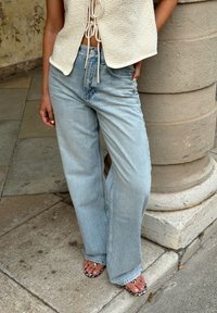 Απαλή μπλε τζιν παντελόνι σε φαρδιά γραμμή με ψηλή μέση και ελαφρώς φθαρμένα σημεία, συνδυασμένα με μαύρες και λευκές σανδάλες με σχέδιο ζώου.