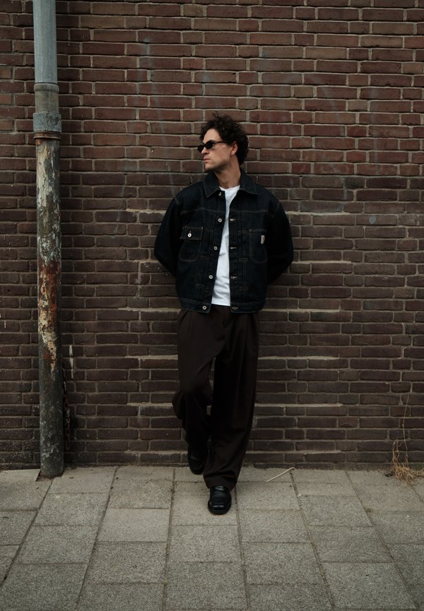 Homme portant des lunettes de soleil, une veste en denim noire, une chemise blanche et un pantalon sombre, appuyé contre un mur de briques marron à côté d’un poteau métallique usé.