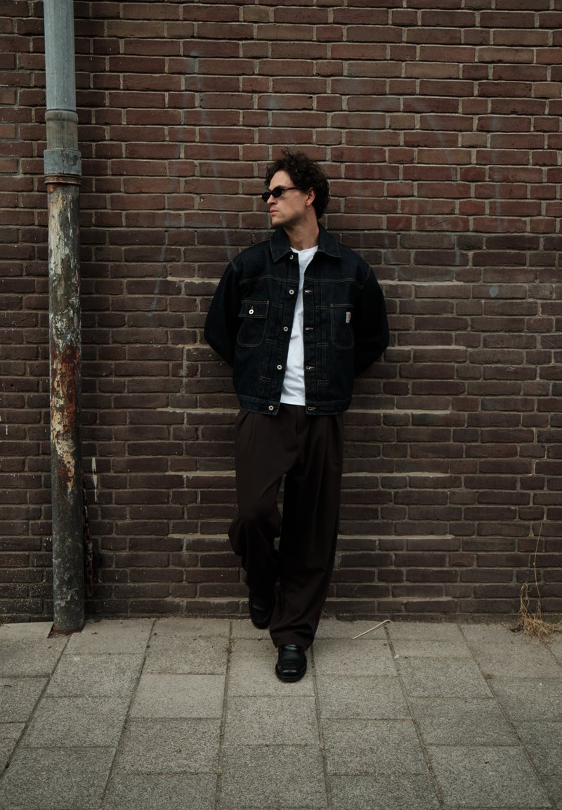 Man wearing sunglasses, black denim jacket, white shirt, and dark pants leaning against a brown brick wall next to a weathered metal pole.