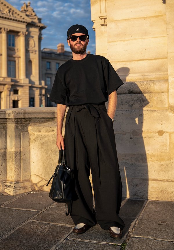 Moški v črnem oversized majici, širokih črnih hlačah, črni kapi, sončnih očalih, ki drži črno usnjeno torbo, stoji ob kamnitem zidu v urbanem okolju.
