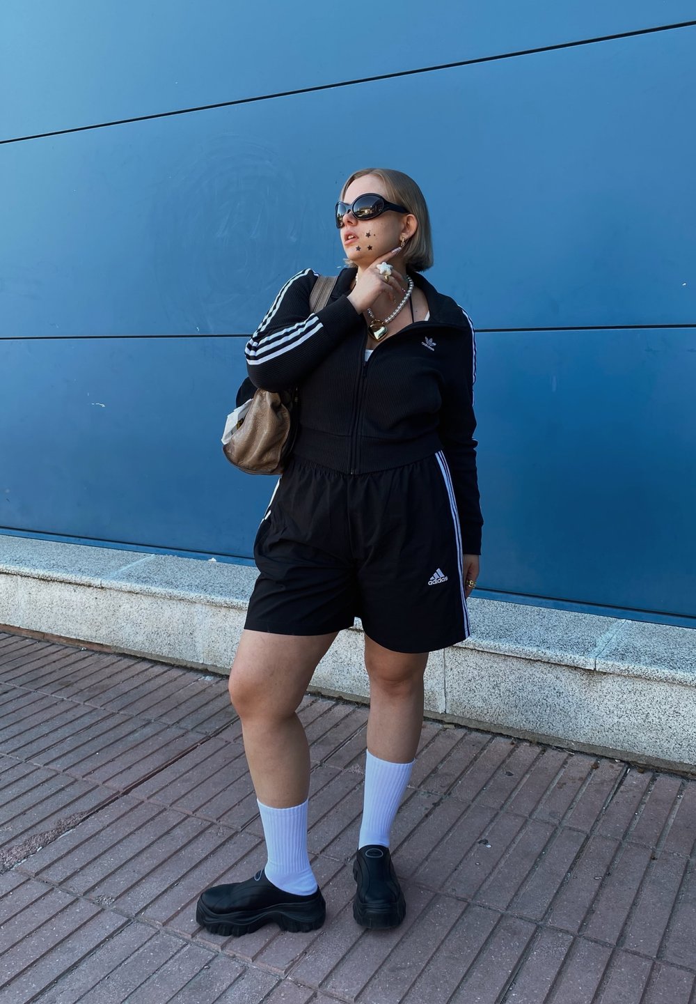 Sort ribbet zip-up jakke og matchende shorts med hvide striber og Adidas-logo. Accessories inkluderer oversized solbriller og en beige taske.