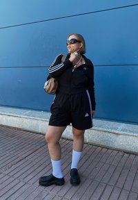 Sort ribbet zip-up jakke og matchende shorts med hvide striber og Adidas-logo. Accessories inkluderer oversized solbriller og en beige taske.