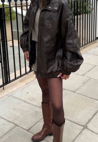 Casaco oversized de pele castanha com punhos em ribana, combinado com um suéter cinza, saia mini preta e botas castanhas até ao joelho.