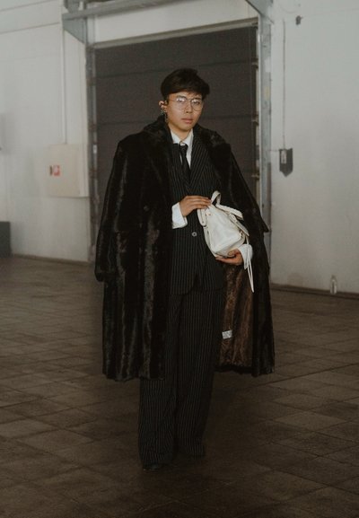 Abrigo de piel sintética negra sobre un traje a rayas, camisa de vestir blanca y sosteniendo un bolso blanco. Las gafas y el auricular son accesorios notables.