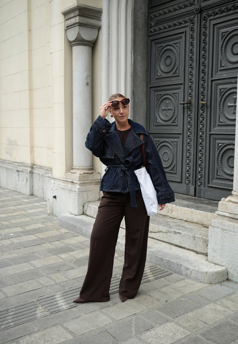 Veste en denim avec taille ceinturée, pantalon long marron et lunettes de soleil. Un sac blanc est porté sur son épaule. Rue en pierre et porte en arrière-plan.