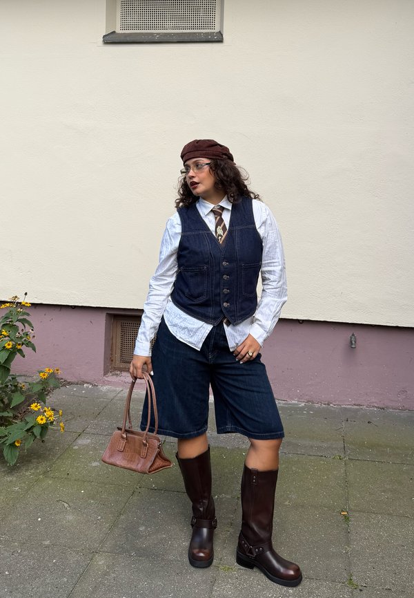 Persona que lleva una boina marrón, gafas, camisa blanca, chaleco oscuro, corbata, pantalones cortos de denim y botas altas marrones, sosteniendo un pequeño bolso marrón.