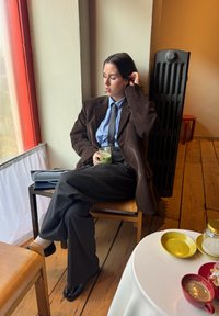 Posedia osoba v tmavohnedom kordovom saku, modrej pruhovanej košeli a sivých nohaviciach, ktorá drží pohár so zeleným nápojom. Teplá drevená podlaha.