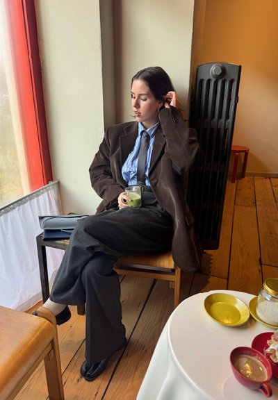 Una persona sentada con un blazer de pana marrón oscuro, camisa azul a rayas y pantalones grises, sosteniendo un vaso con una bebida verde. Suelo cálido de madera.