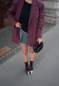 Μπέρντιγγ oversized σακάκι, μαύρο τοπ, γκρι πλισέ φούστα, μαύρη πλεκτή τσάντα, μπέρντιγγ κάλτσες και ασημένια ψηλοτάκουνα παπούτσια με μυτερή μύτη σε στρωμένη επιφάνεια.