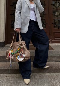 Beige läderhandväska med olika prydnader, med en avslappnad design. Bärs med lösa mörka jeans och en grå blazerkostym med ränder.