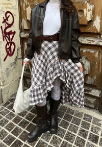 Veste aviateur en cuir marron, col roulé blanc, jupe à carreaux avec des volants, bottes marron jusqu'aux genoux et sac à main blanc. Fond texturé.