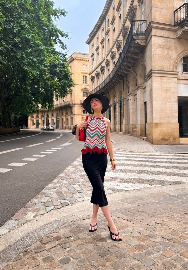 Femeie purtând o pălărie mare neagră, top fără mâneci cu model, fustă neagră și papuci de plajă, mergând pe o stradă pietruită într-o zonă istorică a orașului.