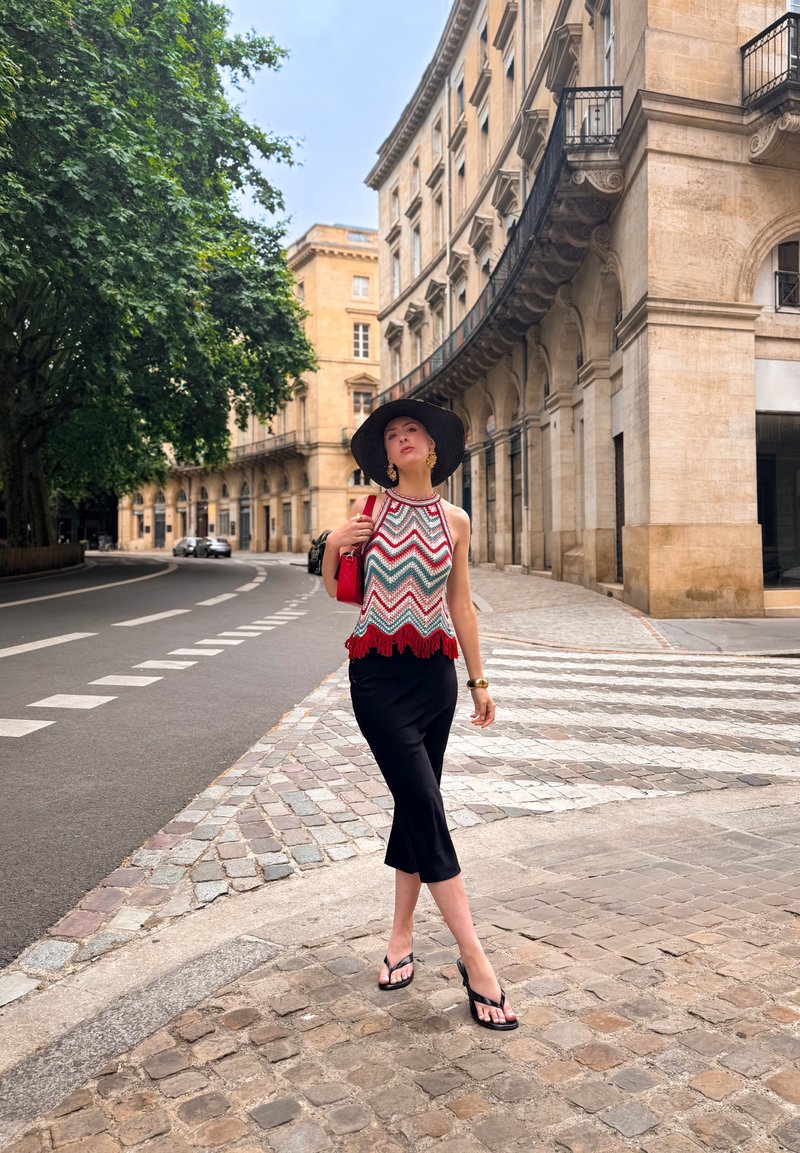 Mujer con un gran sombrero negro, top sin mangas estampado, falda negra y chanclas, caminando por una calle empedrada en una zona histórica de la ciudad.