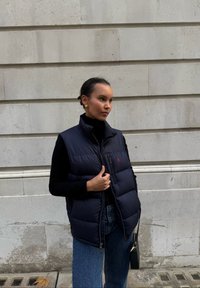 Tumesinine sulejope vest kõrge krae ja tõmblukuga, millel on punane embleem, kombineeritud musta kõrge kaelusega kampsuni ja siniste teksadega.