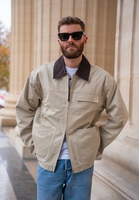 Veste beige en coton avec un col marron, une fermeture éclair frontale et deux grandes poches. Portée sur une chemise blanche et associée à un jean bleu.