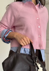 Roze gebreid vest met geribbelde zoom en knoopsluiting, gedragen over een blauw-wit gestreept overhemd met rode accenten, en houdt een bruine leren handtas vast.