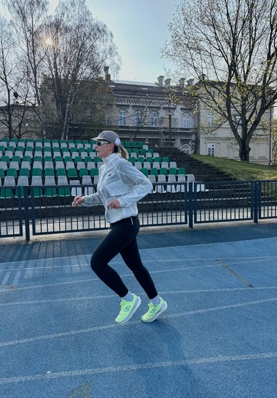 Mujer con chaqueta blanca, mallas negras y zapatillas de neón trotando en una pista azul con gradas vacías y árboles al fondo.