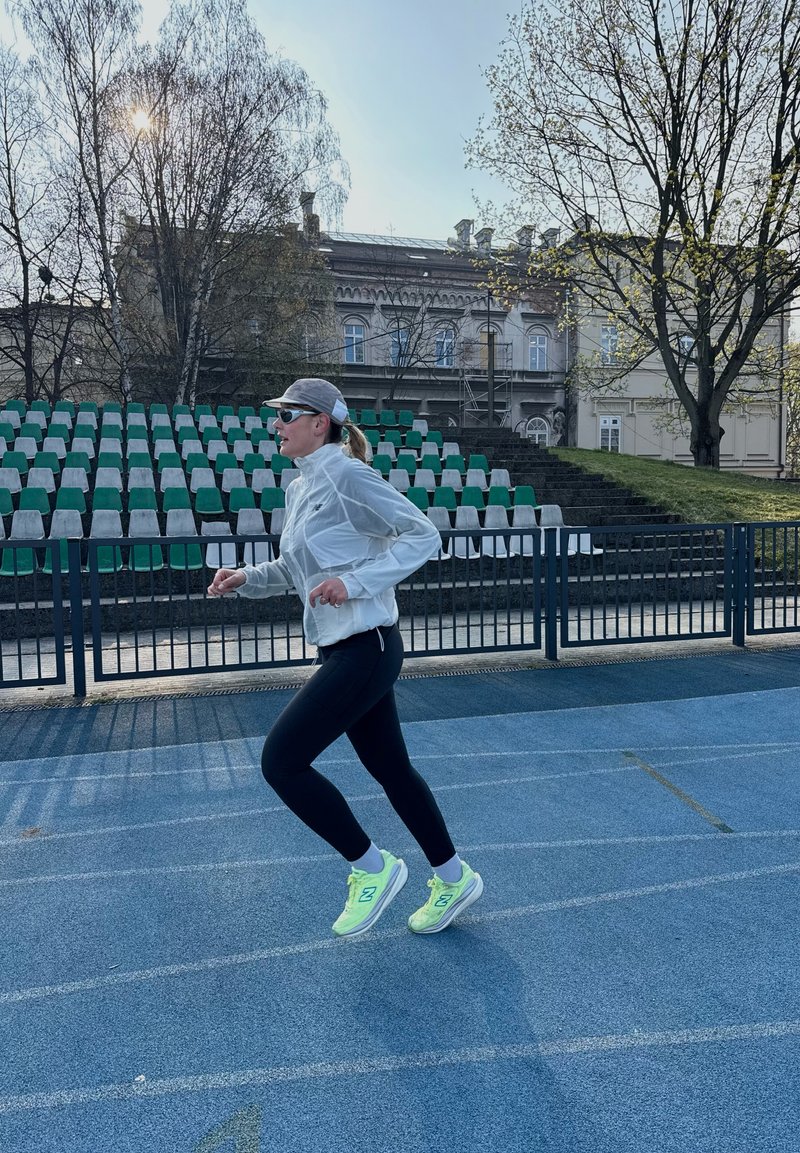 Femme en veste blanche, leggings noirs et baskets néon courant sur une piste bleue avec des gradins vides et des arbres en arrière-plan.