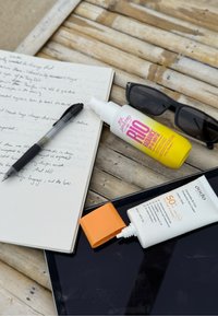 Svart penna, öppen anteckningsbok med handskrivna anteckningar, gul och rosa SPF-sprayflaska, vit solkrämstub med orange kork, svarta solglasögon, surfplatta, träytan.