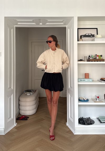 Mujer con gafas de sol, chaqueta beige, falda negra y zapatos rojos, de pie en una puerta en una habitación moderna con estanterías y objetos decorativos.