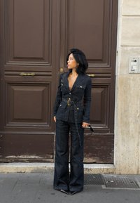 Σκούρο τζιν ολόσωμο με στιλ σακακιού, με χρυσά κουμπιά, ζώνη στη μέση και καμπάνα παντελόνια, φορεμένο με γυαλιά ηλίου.