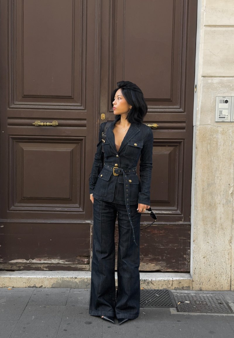 Donker denim jumpsuit met een gestructureerde blazerstijl, voorzien van gouden knopen, een ceintuurlijn en uitlopende broekspijpen, gedragen met een zonnebril.