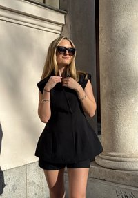 Top negru fără mâneci, cu guler înalt și tiv evazat, asortat cu pantaloni scurți negri. Material neted cu detaliu de fermoar frontal.