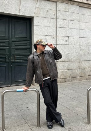 Hombre con chaqueta de cuero marrón, pantalones oscuros, gorra de camuflaje y gafas de sol apoyado en una barandilla de metal frente a un edificio de piedra.