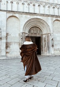 Brązowy szal z imitacji futra o luźnym fasonie, zestawiony z jasnymi dżinsami i czarnymi szpilkami, na tle historycznego kamiennego budynku.